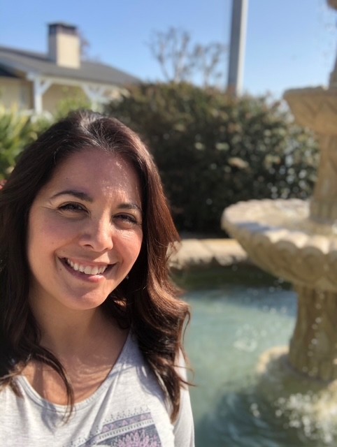 Woman smiling outdoors near a fountain on a sunny day.